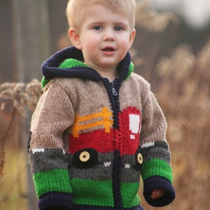 Könnte beinhalten: Ein kleines Kind trägt einen bunten Strickpullover mit einem LKW-Motiv. Der Pullover hat eine Kapuze und ist braun, rot, grün und blau. Das Kind trägt eine blaue Jeans.