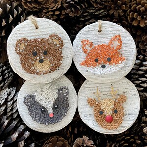 May include: Four round, wooden ornaments with string art animal designs: a bear, fox, badger, and reindeer. Each ornament has a distressed white background and a jute string for hanging. The bear is brown, the fox is orange, the badger is black and white, and the reindeer is tan.