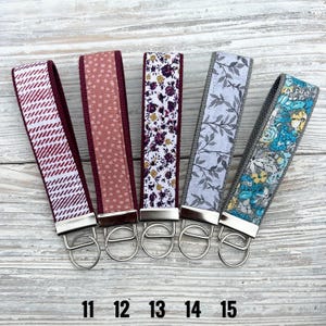 Peut inclure: Cinq porte-cl&eacute;s en tissu avec des accessoires argent&eacute;s. Les porte-cl&eacute;s pr&eacute;sentent divers motifs, dont un chevrons blanc et bordeaux, des pois roses, des motifs floraux, des feuilles grises et un motif floral bleu. Les num&eacute;ros 11, 12, 13, 14 et 15 sont imprim&eacute;s en bas.