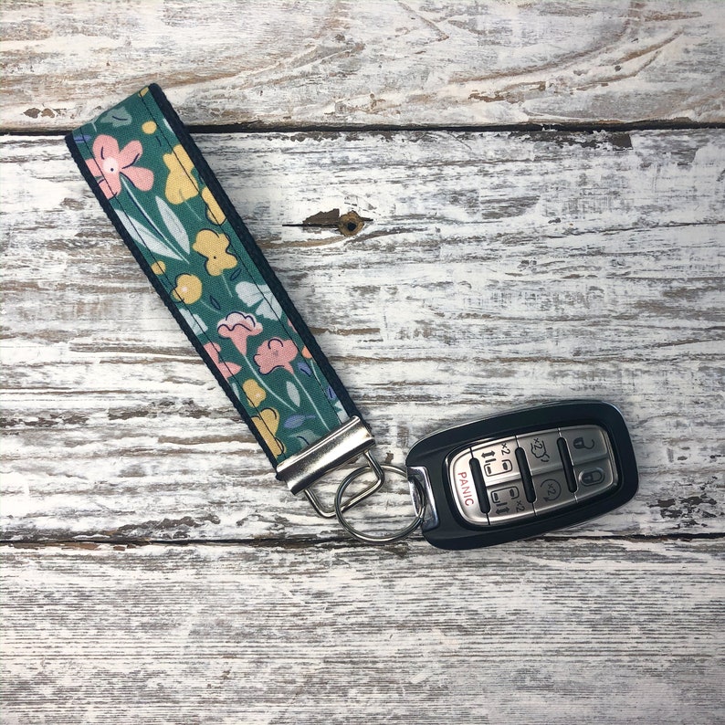 Peut inclure: Un porte-cl&eacute;s floral vert d'eau avec des fleurs roses et jaunes, des feuilles blanches et une bordure noire. Il est attach&eacute; &agrave; un porte-cl&eacute;s argent&eacute; et &agrave; une cl&eacute; de voiture noire avec des boutons.