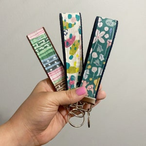 Peut inclure: Trois porte-cl&eacute;s en tissu avec diff&eacute;rents motifs sont tenus dans une main. Les motifs comprennent un design ray&eacute; rose et vert, un design abstrait color&eacute; et un motif floral. Chaque porte-cl&eacute;s a une bordure bleu fonc&eacute; et un porte-cl&eacute;s argent&eacute;.