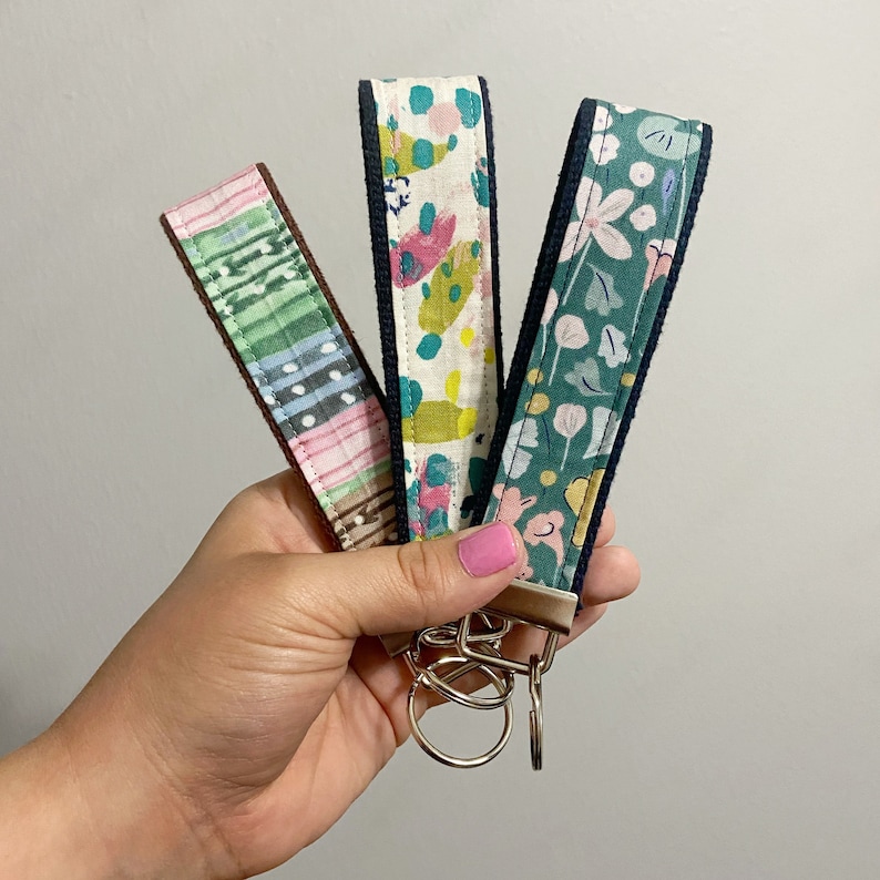 Peut inclure: Trois porte-cl&eacute;s en tissu avec diff&eacute;rents motifs sont tenus dans une main. Les motifs comprennent un design ray&eacute; rose et vert, un design abstrait color&eacute; et un motif floral. Chaque porte-cl&eacute;s a une bordure bleu fonc&eacute; et un porte-cl&eacute;s argent&eacute;.