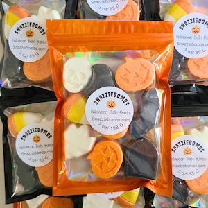 May include: A clear, iridescent-edged pouch containing assorted Halloween-themed bath bombs in shapes of white skulls, orange pumpkins, black bats, and candy corn. The label reads SNAZZIEBOMBS, Halloween Bath Bombs, Snazziebombs.com, 7 oz/ 198 G.