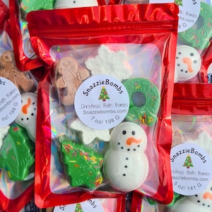 May include: A red holographic pouch containing assorted Christmas-themed bath bombs, including a brown gingerbread man, white snowflakes, a green wreath, a green tree, and a white snowman. A label reads SnazzieBombs Christmas Bath Bombs 7 oz/ 198 G.