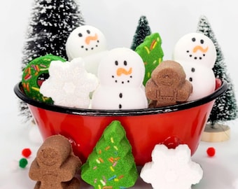 May include: A red bowl filled with assorted holiday-themed bath bombs, including white snowmen with orange noses, brown gingerbread figures, green Christmas trees with colorful sprinkles, and white snowflakes.