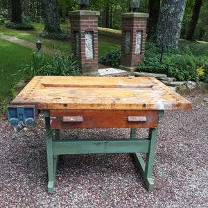 May include: A rectangular wooden workbench featuring a thick, worn natural wood top with a central recessed channel and a series of holes along the edges. It includes a blue metal vise on the left side and a single wooden drawer with two handles.