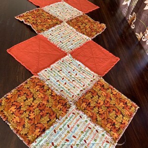 May include: A patchwork table runner with a fall theme. The runner is made of different fabrics with various patterns, including leaves, words, and a solid orange color. The runner has a frayed edge.