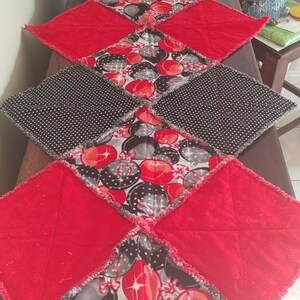 May include: A red and black quilted table runner with a pattern of red and white ornaments. The runner is made of fabric squares sewn together with a frayed edge.