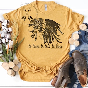 Op de afbeelding: Een mosterdgeel T-shirt met een zwart-witte afbeelding van een indiaanse vrouw met een hoofdtooi. De tekst "Be Brave, Be Bold, Be Fierce" is onder de afbeelding gedrukt. Het shirt ligt plat op een wit houten oppervlak.