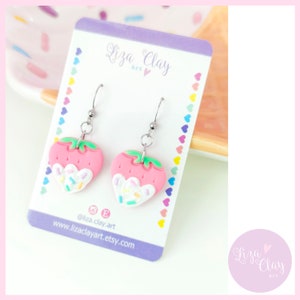 Puede incluir: Un par de pendientes colgantes con forma de fresa rosa con glaseado blanco y chispas de colores. Los pendientes están hechos de arcilla polimérica y tienen ganchos plateados. Los pendientes se muestran en una tarjeta blanca con el texto "Liza Clay Art" y el nombre de usuario de las redes sociales "@liza.clay.art".