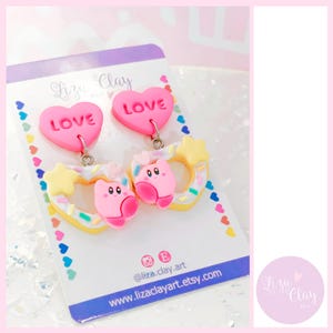 Puede incluir: Pendientes de arcilla polimérica rosa con un corazón con la palabra "LOVE" y una dona con un personaje rosa y espolvoreado. Los pendientes están en una tarjeta blanca con el texto "Liza Clay Art" y el nombre de usuario de las redes sociales "@liza.clay.art".