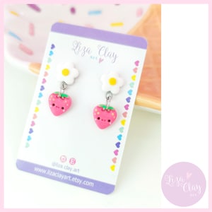 Puede incluir: Un par de pendientes colgantes con una fresa rosa y un dije de margarita blanca. Los pendientes están en una tarjeta morada con el texto "Liza Clay Art" y la dirección del sitio web "www.lizaclayart.etsy.com".