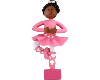 May include: A pink ballerina figurine with dark skin, wearing a pink leotard, tutu, and ballet shoes. The figurine has a flower in her hair and is posed en pointe with arms crossed.