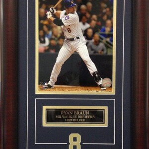 May include: Framed photo of baseball player Ryan Braun of the Milwaukee Brewers. The image shows Braun in a batting stance, with a baseball in the foreground. The frame includes a nameplate and the number 8.