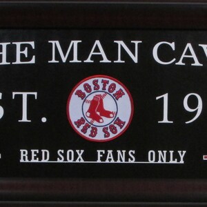 Puede incluir: Un letrero negro enmarcado con texto blanco que dice "The Man Cave Est. 1901 Red Sox Fans Only". Un logotipo de los Red Sox de Boston rojo, blanco y azul está en el centro.