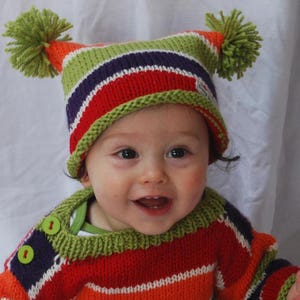 May include: A child wearing a striped orange, green, and purple knitted hat with green pom-poms and a striped orange, green, and purple knitted sweater.