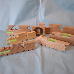 May include: A collection of light-coloured wooden train-shaped bottle openers, each featuring a small horizontal spirit level with green liquid. One piece includes a circular metal bottle opener insert, while the others show the wood grain.