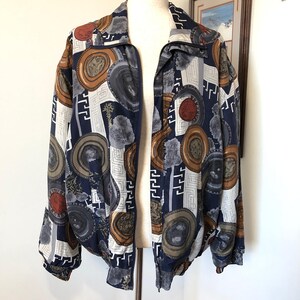 May include: A vintage bomber jacket with a full zipper, displayed on a mannequin. The jacket features a navy blue background with a repeating pattern of circular and geometric shapes in shades of brown, gray, and white. The jacket has a ribbed cuff and hem.