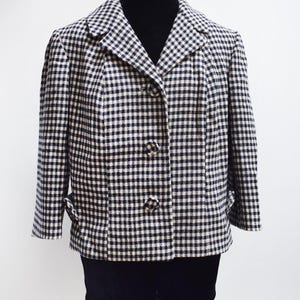 Könnte beinhalten: Ein schwarz-weiß karierter Blazer mit klassischem Kragen und drei passenden Knöpfen. Der Blazer hat zwei Vordertaschen und wird auf einer schwarzen Schaufensterpuppe präsentiert. Das Muster ist ein kleiner Vichy-Karo.