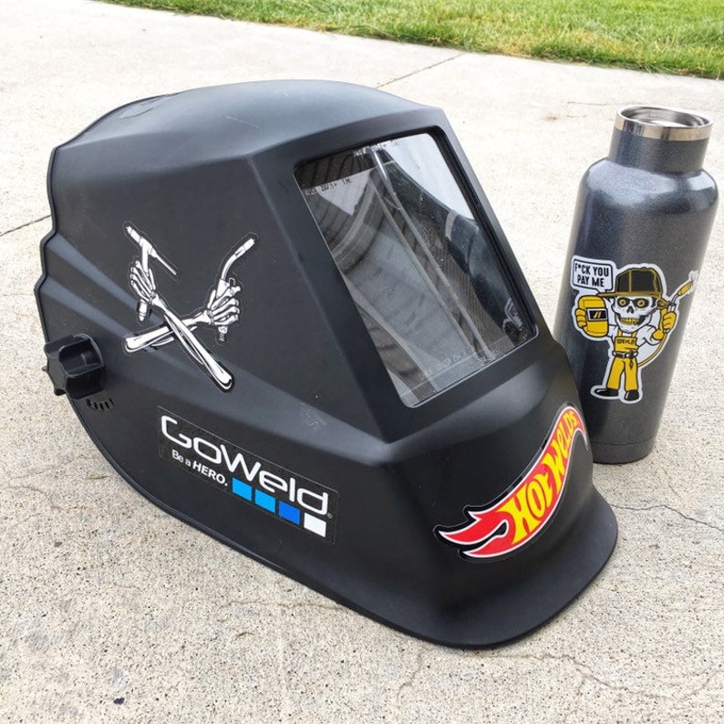 Op de afbeelding: Een zwarte lashelm met een helder vizier. De helm heeft verschillende stickers, waaronder een skelet dat lasgereedschap vasthoudt, een Hot Wheels-logo en een GoPro-logo. De helm staat op een grijs betonnen oppervlak.