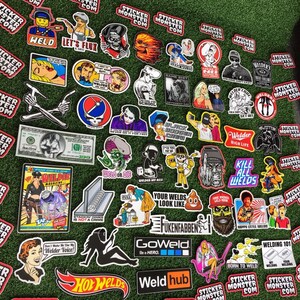 Op de afbeelding: Een verzameling kleurrijke stickers met verschillende ontwerpen, waaronder een schedel, een lasser, een hotrod en een cartoonfiguur. De stickers zijn gerangschikt op een groene ondergrond. De tekst "Sticker Monster" staat op verschillende stickers.