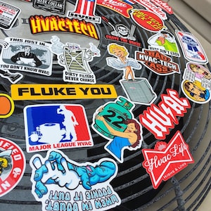 May include: A collection of colorful stickers with various designs and text, including "HVAC Tech", "Fluke You", "Major League HVAC", and "Hvac Life". The stickers are arranged on a metal surface with a grid pattern.