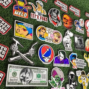 Op de afbeelding: Een verzameling kleurrijke stickers met verschillende ontwerpen, waaronder een schedel, een lasser, een cartoonfiguur en een afbeelding van een band. De stickers bevinden zich op een groene achtergrond en bevatten tekst zoals "Sticker Monster", "Weld", "Let's Flux", "Too Cool for School" en "Same Shit Different Day".