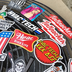 May include: A collection of colorful stickers with various designs, including a red, white, and blue American flag, a red and yellow sticker with the text "HVAC TECHS KICK ASS EVERY DAMN DAY", a red and white sticker with the text "HVAC TECH - A REAL AMERICAN HERO", and a red and white sticker with the text "Huac HIGH LIFE".