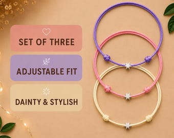 Pode incluir: Um conjunto de três pulseiras ajustáveis com cordões roxos, rosa e creme, cada uma centrada com um pequeno pingente de estrela em tom prateado. O texto na imagem diz: SET OF THREE, ADJUSTABLE FIT, DAINTY & STYLISH.