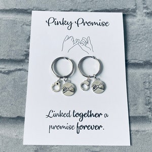 May include: Two silver keychains with a pinky promise design. The keychains are linked together with a small silver charm. The card reads "Pinky Promise" and "Linked together a promise forever."