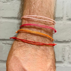 May include: A person's wrist with four colourful string bracelets. The bracelets are layered in a gradient from pink to red.