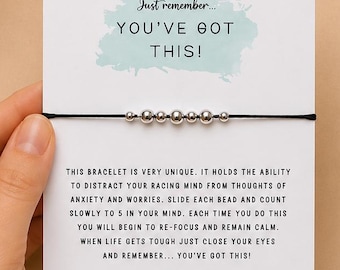 May include: A thin black cord bracelet featuring five small silver-toned metallic beads, mounted on a white display card. The card displays the text "Just remember... YOU'VE GOT THIS!" and instructions for using the beads to focus.