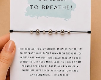 May include: A thin black cord bracelet featuring seven silver-toned beads of varying sizes, mounted on a white display card. The card includes the text Just remember... TO BREATHE! and instructions for using the beads to focus.