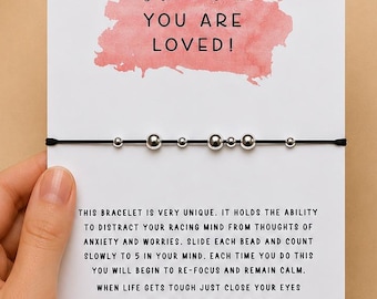 May include: A black cord bracelet with silver-toned beads attached to a white display card. The card features a pink watercolor splash with the text: Just remember... YOU ARE LOVED! and instructions for using the beads to focus and remain calm.