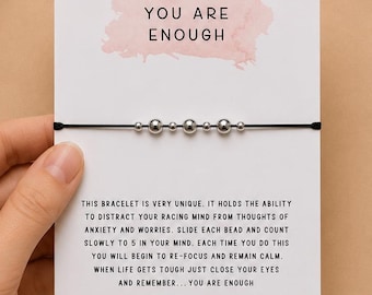 May include: A black cord bracelet with five silver-toned beads attached to a white display card. The card features a pink watercolor accent and text that reads: Just remember... YOU ARE ENOUGH, followed by instructions for use and a website URL.