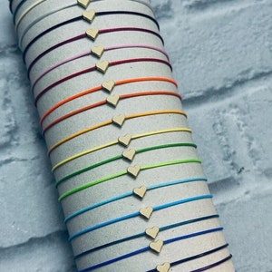 Könnte beinhalten: Eine Sammlung bunter Armbänder aus Schnur mit Holzherzen als Anhänger. Die Armbänder sind in einem Stapel angeordnet, wobei die Farben von Grau bis Lila reichen.