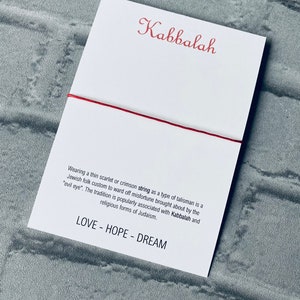Pode incluir: Um bracelete de cord&atilde;o vermelho com a palavra "Kabbalah" escrita em it&aacute;lico em um cart&atilde;o branco. O cart&atilde;o tamb&eacute;m inclui texto sobre a tradi&ccedil;&atilde;o de usar um cord&atilde;o vermelho como talism&atilde; para evitar a m&aacute; sorte. O texto diz "Usar um cord&atilde;o fino escarlate ou carmesim como tipo de talism&atilde; &eacute; um costume popular judeu para evitar a m&aacute; sorte causada pelo "mau-olhado". A tradi&ccedil;&atilde;o &eacute; popularmente associada &agrave; Cabala e &agrave;s formas religiosas do juda&iacute;smo. LOVE - HOPE - DREAM"