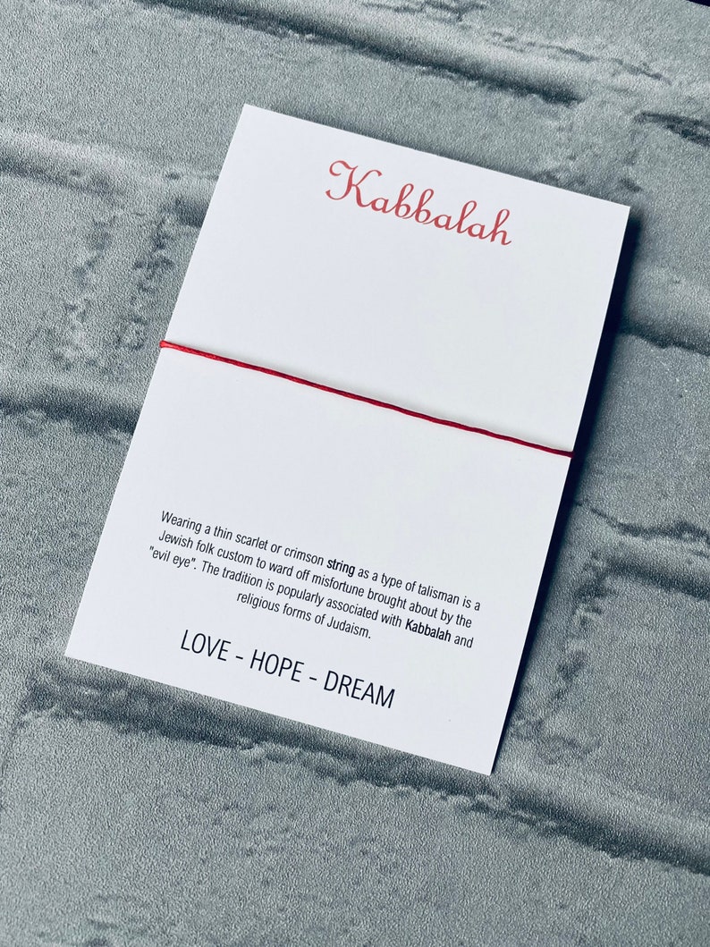 Pode incluir: Um bracelete de cord&atilde;o vermelho com a palavra "Kabbalah" escrita em it&aacute;lico em um cart&atilde;o branco. O cart&atilde;o tamb&eacute;m inclui texto sobre a tradi&ccedil;&atilde;o de usar um cord&atilde;o vermelho como talism&atilde; para evitar a m&aacute; sorte. O texto diz "Usar um cord&atilde;o fino escarlate ou carmesim como tipo de talism&atilde; &eacute; um costume popular judeu para evitar a m&aacute; sorte causada pelo "mau-olhado". A tradi&ccedil;&atilde;o &eacute; popularmente associada &agrave; Cabala e &agrave;s formas religiosas do juda&iacute;smo. LOVE - HOPE - DREAM"