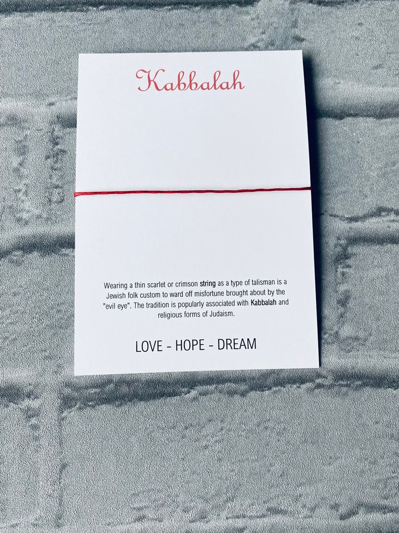 Pode incluir: Um bracelete de cord&atilde;o vermelho com a palavra "Kabbalah" escrita em it&aacute;lico em um cart&atilde;o branco. O cart&atilde;o tamb&eacute;m inclui texto sobre o significado do cord&atilde;o na tradi&ccedil;&atilde;o judaica e as palavras "LOVE - HOPE - DREAM".
