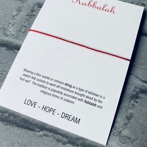 Pode incluir: Um cord&atilde;o vermelho amarrado em torno de um cart&atilde;o branco com a palavra "Kabbalah" escrita em it&aacute;lico vermelho. O cart&atilde;o tem texto sobre o significado do cord&atilde;o na tradi&ccedil;&atilde;o judaica e as palavras "LOVE - HOPE - DREAM" impressas na parte inferior.