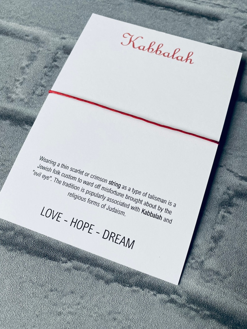 Pode incluir: Um cord&atilde;o vermelho amarrado em torno de um cart&atilde;o branco com a palavra "Kabbalah" escrita em it&aacute;lico vermelho. O cart&atilde;o tem texto sobre o significado do cord&atilde;o na tradi&ccedil;&atilde;o judaica e as palavras "LOVE - HOPE - DREAM" impressas na parte inferior.