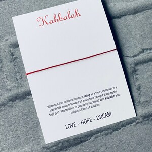 Pode incluir: Um bracelete de corda vermelha com a palavra "Kabbalah" escrita em it&aacute;lico vermelho em um cart&atilde;o branco. O cart&atilde;o tamb&eacute;m inclui o texto "Usar um fio fino escarlate ou carmesim como um tipo de talism&atilde; &eacute; um costume popular judeu para evitar a m&aacute; sorte causada pelo "mau-olhado". A tradi&ccedil;&atilde;o &eacute; popularmente associada &agrave; Cabala e &agrave;s formas religiosas do juda&iacute;smo." e "LOVE - HOPE - DREAM".