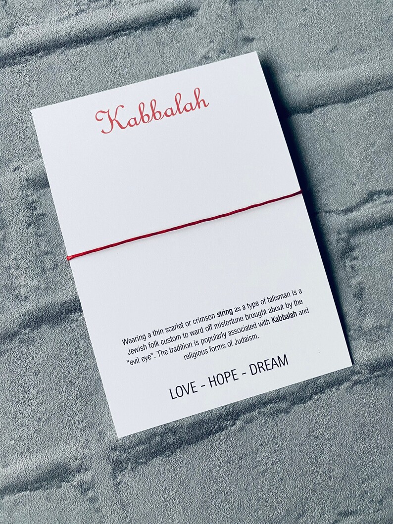 Pode incluir: Um bracelete de corda vermelha com a palavra "Kabbalah" escrita em it&aacute;lico vermelho em um cart&atilde;o branco. O cart&atilde;o tamb&eacute;m inclui o texto "Usar um fio fino escarlate ou carmesim como um tipo de talism&atilde; &eacute; um costume popular judeu para evitar a m&aacute; sorte causada pelo "mau-olhado". A tradi&ccedil;&atilde;o &eacute; popularmente associada &agrave; Cabala e &agrave;s formas religiosas do juda&iacute;smo." e "LOVE - HOPE - DREAM".