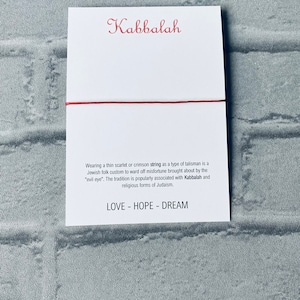 Pode incluir: Um fio vermelho amarrado em torno de um cart&atilde;o branco com a palavra "Kabbalah" escrita em it&aacute;lico vermelho. O cart&atilde;o tamb&eacute;m tem texto sobre o significado do fio na tradi&ccedil;&atilde;o judaica e as palavras "LOVE - HOPE - DREAM" impressas nele.