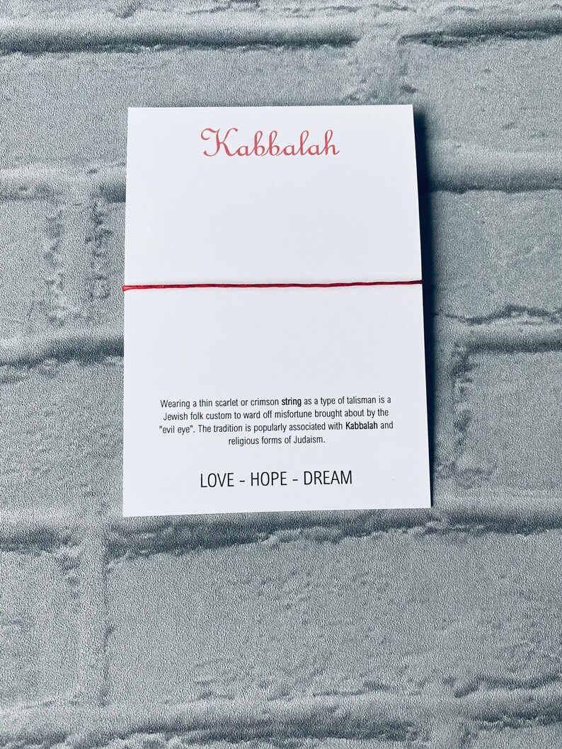 Pode incluir: Um fio vermelho amarrado em torno de um cart&atilde;o branco com a palavra "Kabbalah" escrita em it&aacute;lico vermelho. O cart&atilde;o tamb&eacute;m tem texto sobre o significado do fio na tradi&ccedil;&atilde;o judaica e as palavras "LOVE - HOPE - DREAM" impressas nele.