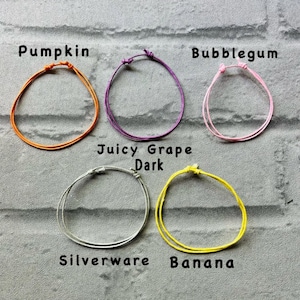 May include: Five colourful friendship bracelets with different names written below each one: Pumpkin, Bubblegum, Juicy Grape Dark, Silverware, and Banana.