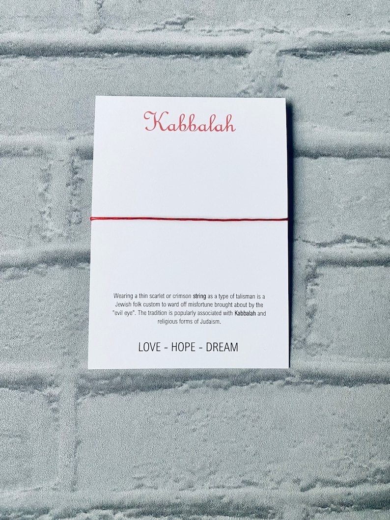 Pode incluir: Um bracelete de cord&atilde;o vermelho com a palavra "Kabbalah" e a frase "LOVE - HOPE - DREAM" impressa em um cart&atilde;o branco.