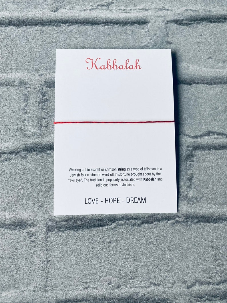 Pode incluir: Um bracelete de cord&atilde;o vermelho com a palavra "Kabbalah" escrita em it&aacute;lico em um cart&atilde;o branco. O cart&atilde;o tamb&eacute;m inclui texto sobre a tradi&ccedil;&atilde;o de usar um cord&atilde;o vermelho como talism&atilde; para evitar a m&aacute; sorte. O texto tamb&eacute;m menciona a associa&ccedil;&atilde;o com a Cabala e as formas religiosas do juda&iacute;smo. O cart&atilde;o tamb&eacute;m inclui as palavras "LOVE - HOPE - DREAM".