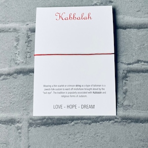 Pode incluir: Um bracelete de cord&atilde;o vermelho com a palavra "Kabbalah" impressa em um cart&atilde;o branco. O cart&atilde;o tamb&eacute;m inclui texto sobre a tradi&ccedil;&atilde;o de usar um cord&atilde;o vermelho como talism&atilde; para evitar a m&aacute; sorte.
