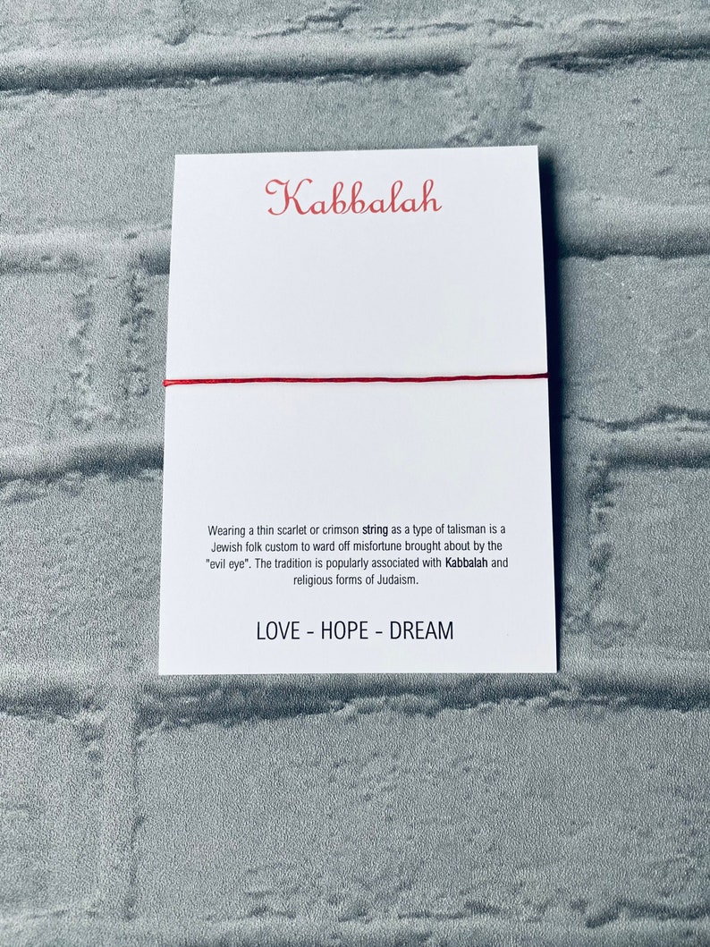 Pode incluir: Um bracelete de cord&atilde;o vermelho com a palavra "Kabbalah" impressa em um cart&atilde;o branco. O cart&atilde;o tamb&eacute;m inclui texto sobre a tradi&ccedil;&atilde;o de usar um cord&atilde;o vermelho como talism&atilde; para evitar a m&aacute; sorte.